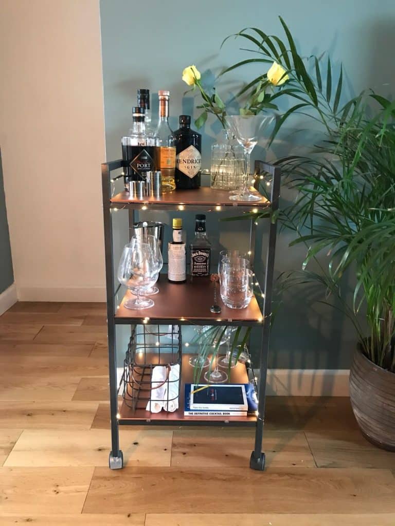 Three-tier copper and black bar cart styled with fairy lights, spirits, glassware, and a vase of yellow roses against a teal wall