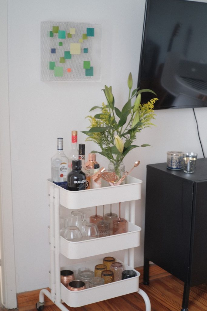 White three-tier utility cart styled with fresh lily and wildflower stems, copper cocktail tools, and glassware next to a black dresser