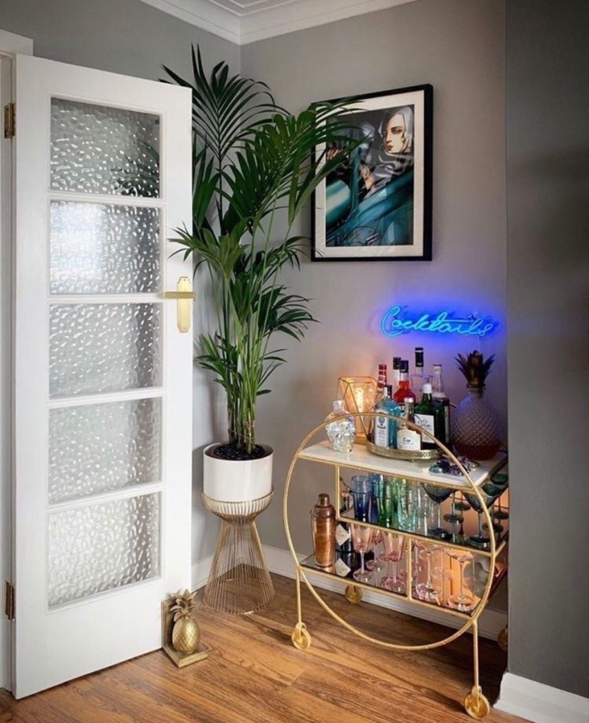 Round gold bar cart with mirrored shelves styled with colourful glasses, candle holders, and spirits beneath a blue neon cocktails sign and a framed art print in a grey room with a large palm plant