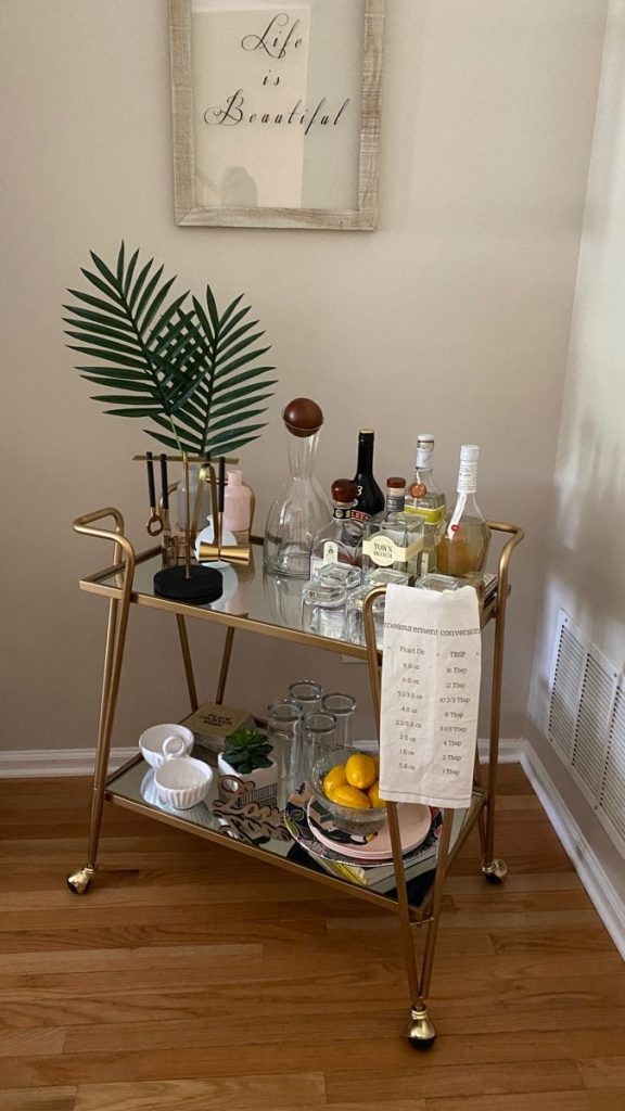 Gold two-tier mirrored bar cart styled with a faux palm stem, gold bar tools, clear decanters, and fresh lemons beneath a Life is Beautiful framed print