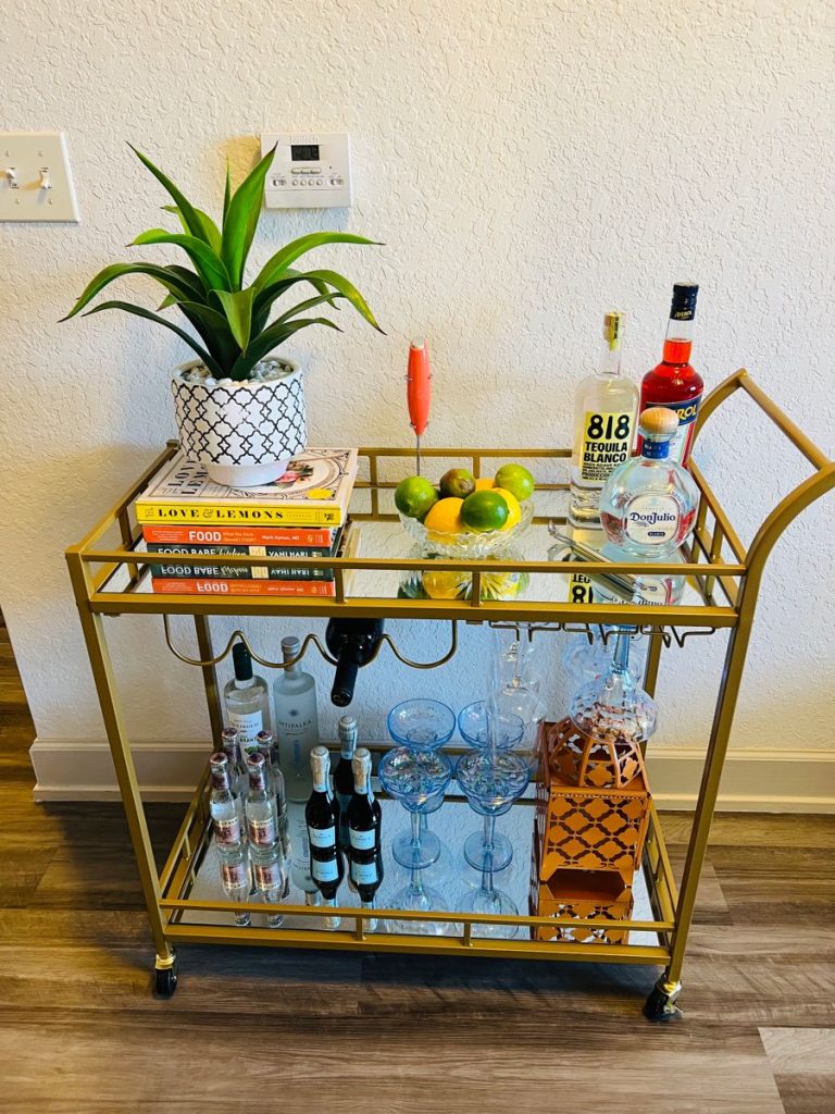 Gold three-tier bar cart with built-in wine rack and glass hangers, styled with colourful cookbooks, fresh citrus in a bowl, and 818 Tequila bottles on top