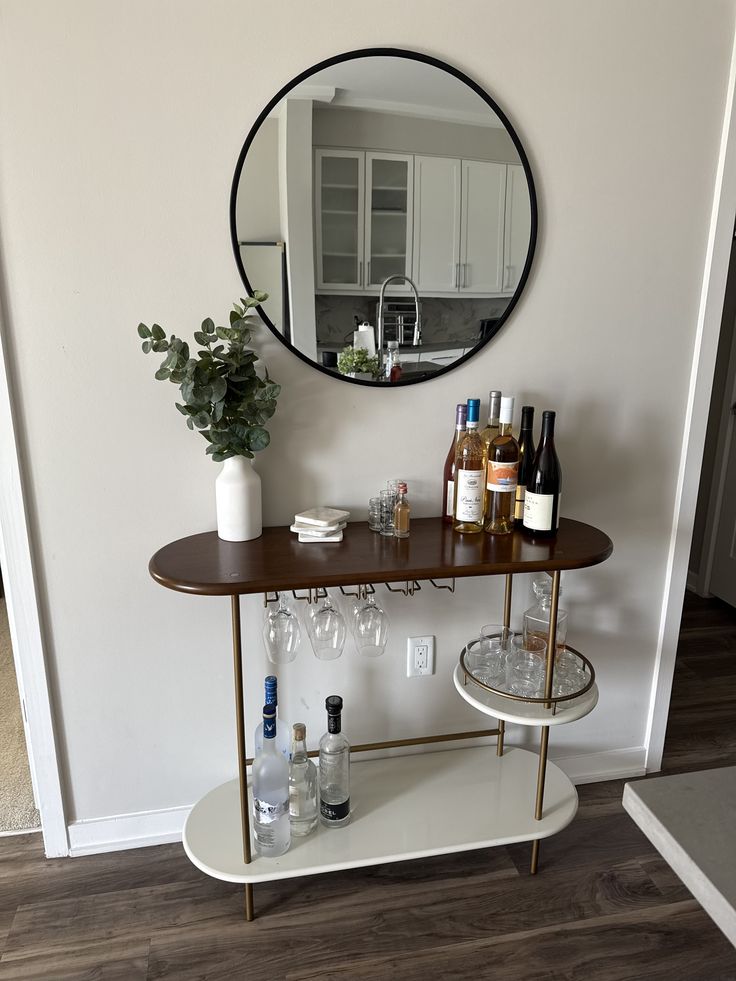 Two-tier walnut and gold bar table with hanging wine glass rack, a white vase of eucalyptus stems, wine bottles, and a round black-framed mirror above in a neutral room