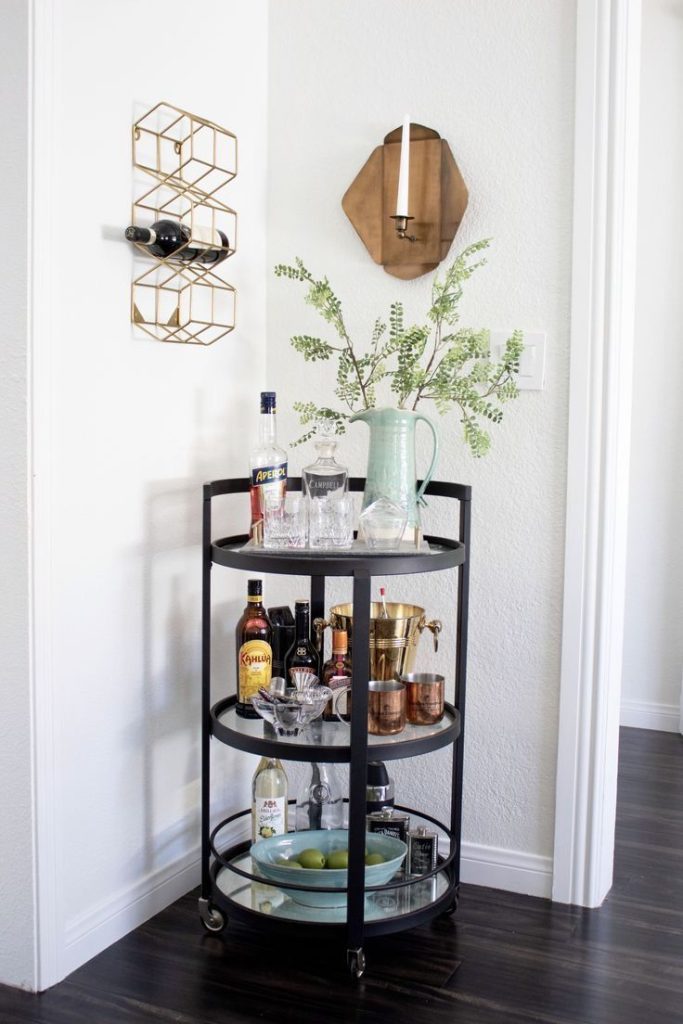 Black three-tier round bar cart styled with a mint ceramic jug of greenery, crystal decanters, and copper mugs beside a gold geometric wall wine rack and wooden wall sconce
