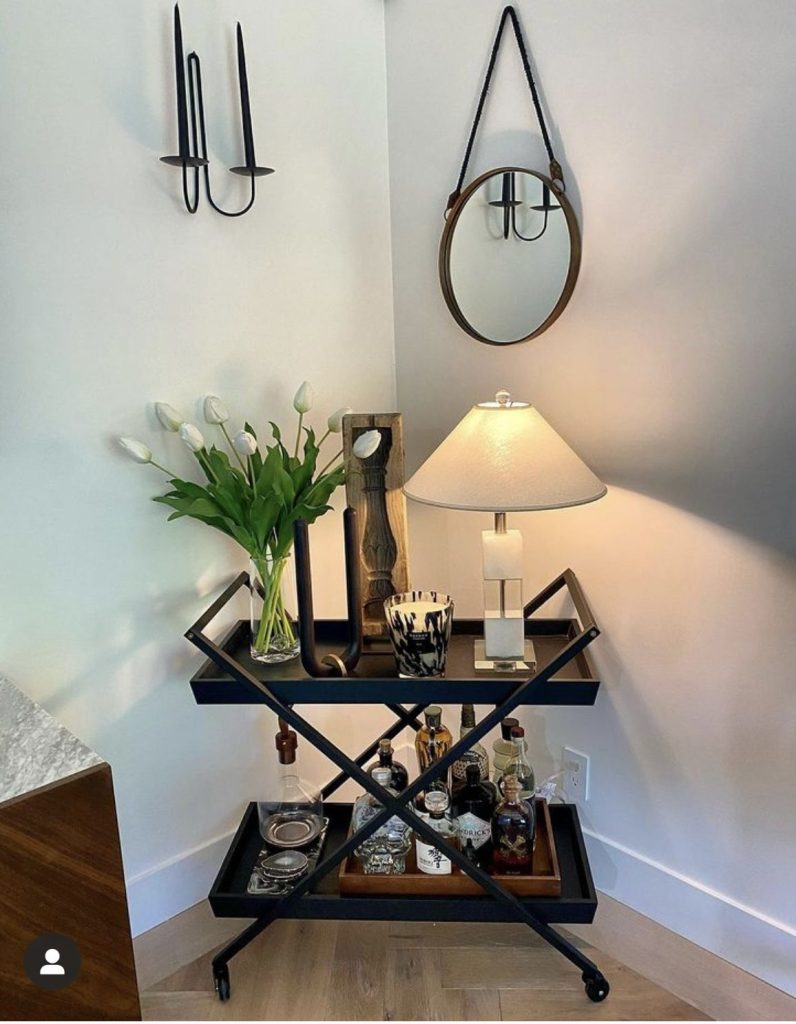 Black X-frame bar cart styled with white tulips, a table lamp, a tortoiseshell candle, and spirits on the lower shelf in a corner with a round leather-strap mirror and black wall sconces above