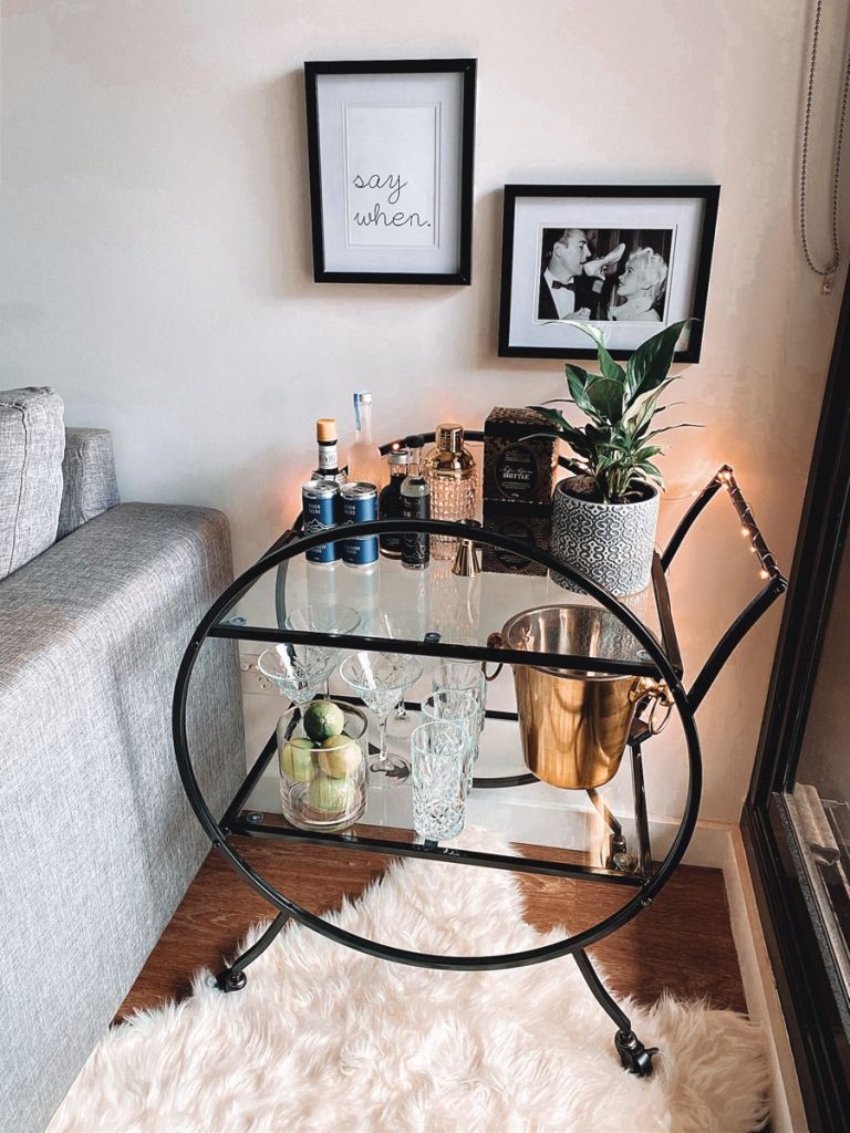 Black oval two-tier bar cart with glass shelves styled with a gold ice bucket, copper shaker, crystal glasses, and a small plant beside a grey sofa with say when and vintage Hollywood framed prints above