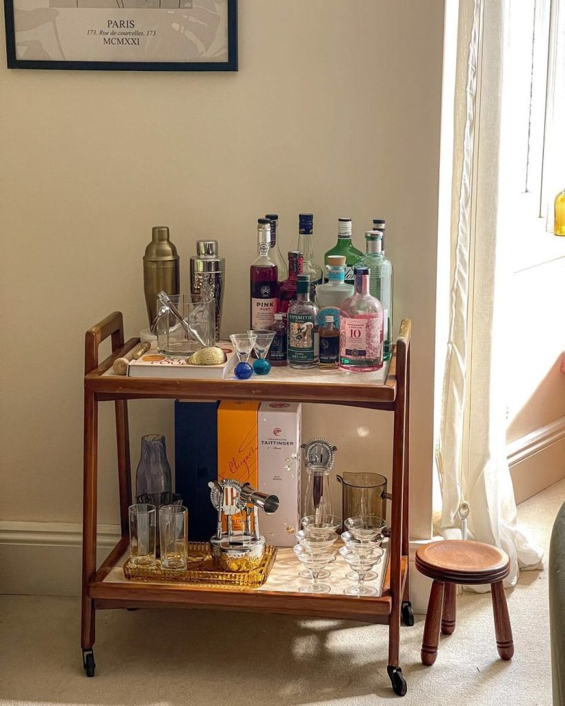 Wooden two-tier bar cart packed with colourful gin bottles, cocktail shakers, and glassware near a bright window with a Paris print above