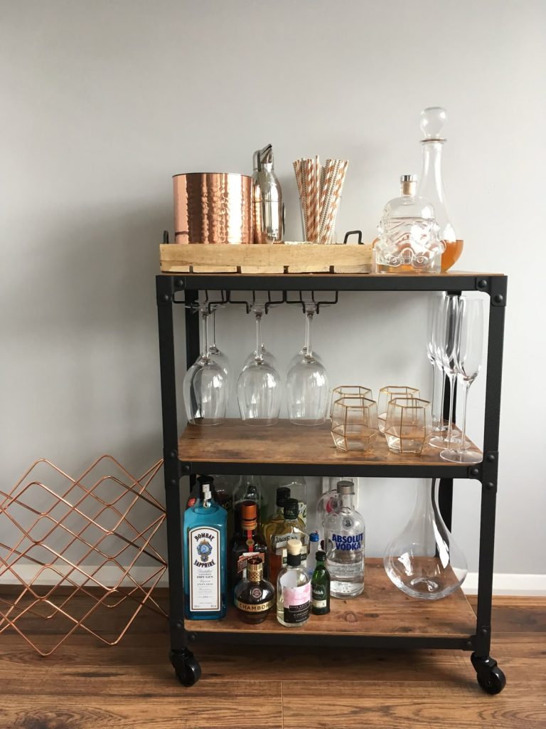 Black and wood three-tier industrial bar cart with hanging wine glasses, a copper ice bucket on a wooden tray, a crystal decanter, and gold geometric glasses with a copper wine rack beside it
