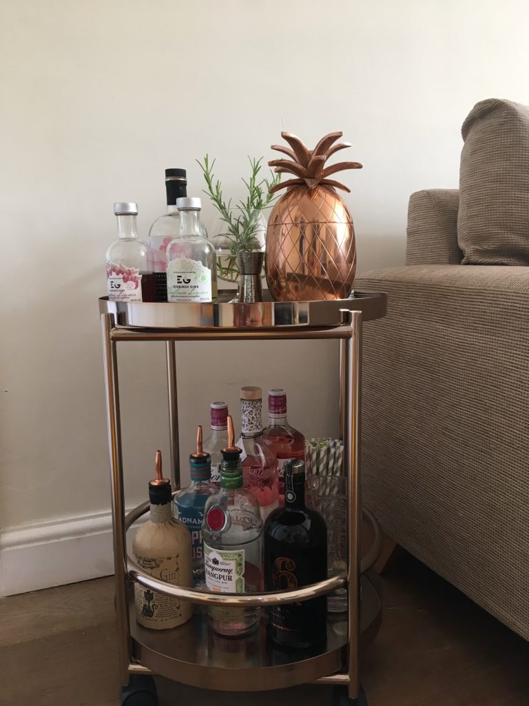 Gold and glass two-tier round bar cart styled with a copper pineapple accent piece, fresh rosemary in a small vase, and a collection of Edinburgh Gin bottles next to a sofa