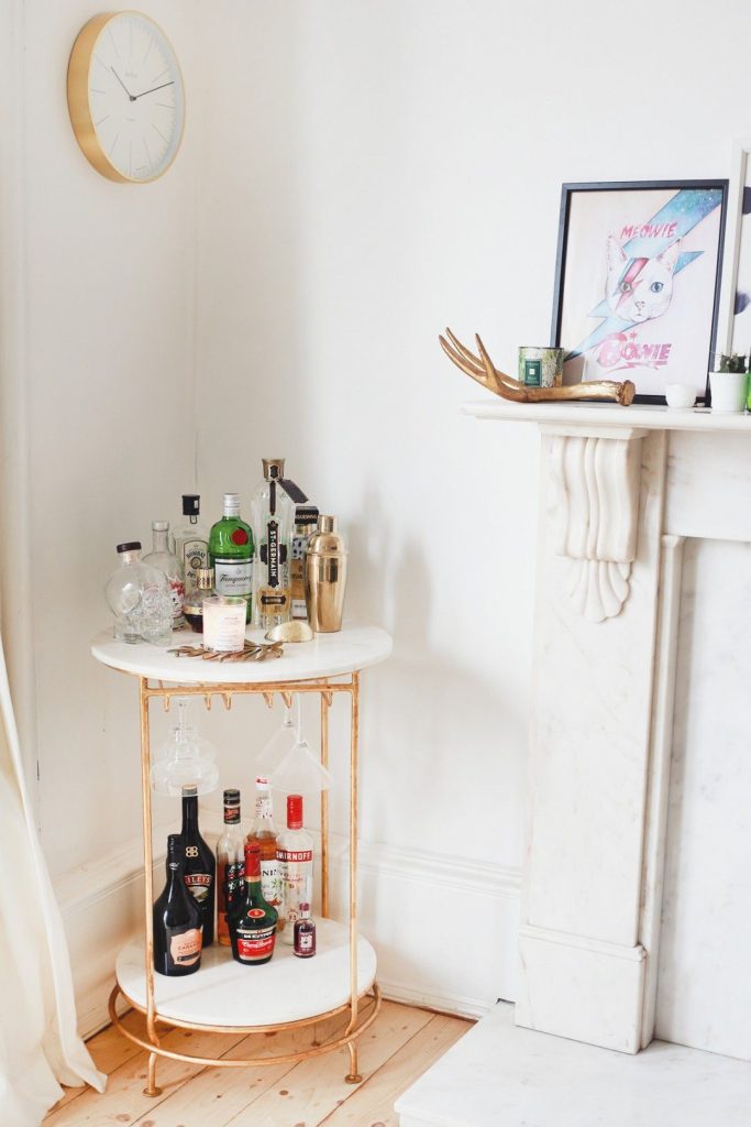 Round white marble and gold two-tier bar cart with hanging wine glasses and a collection of spirits beside a white marble fireplace with a gold shaker and a scented candle on top