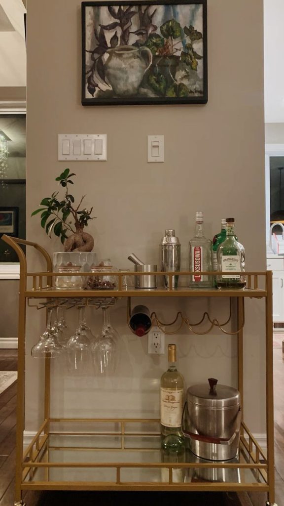 Gold and mirrored two-tier bar cart with built-in wine rack and hanging wine glasses, styled with a small bonsai plant, cocktail shakers, and spirits in an open-plan living room