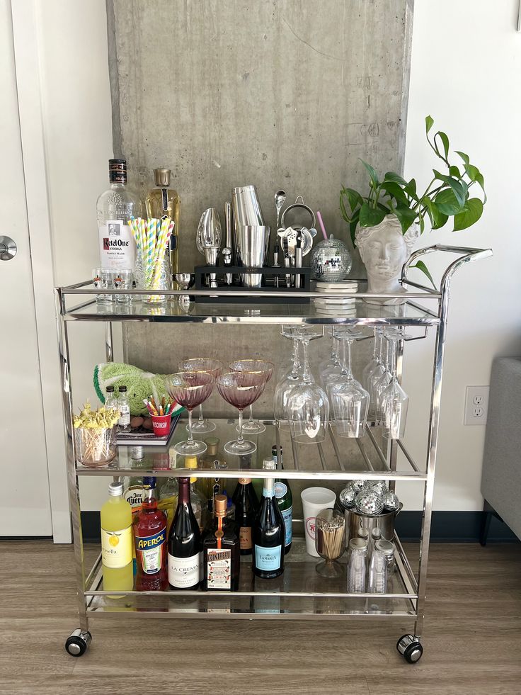 Silver chrome three-tier bar cart with glass shelves styled with a sculptural head planter with a trailing plant, pink coupe glasses, a mini disco ball, and a full collection of spirits and mixers