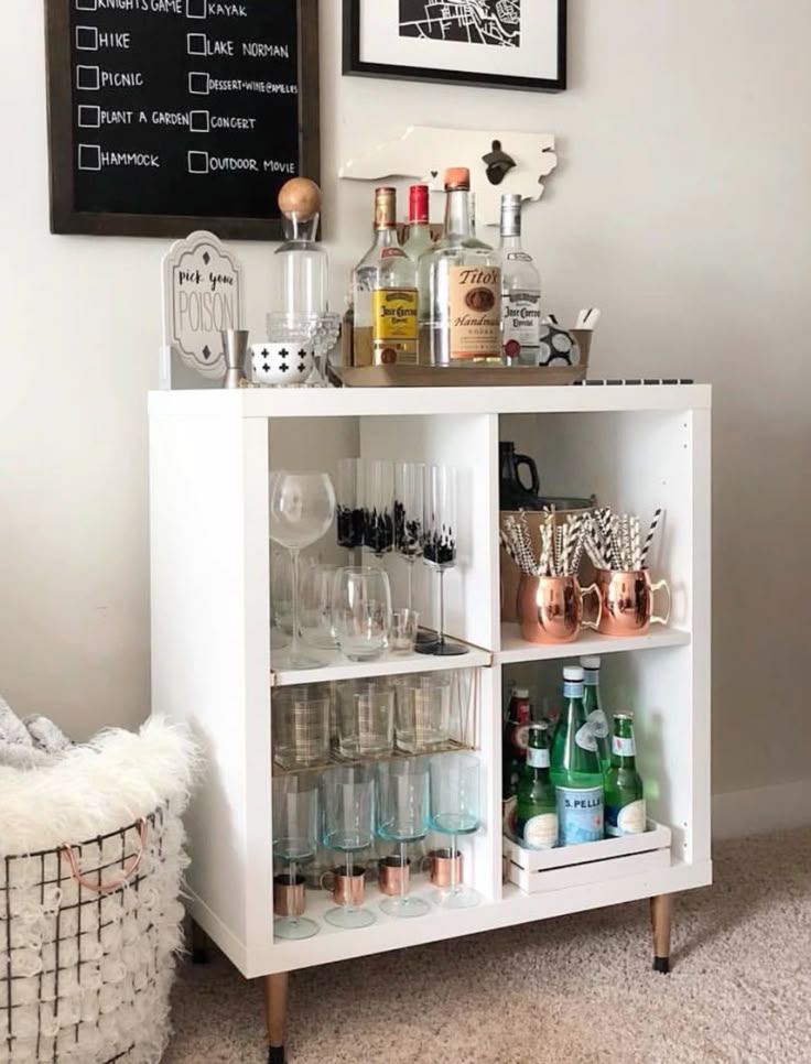 White four-cubby bookcase with mid-century legs styled as a home bar with wine glasses, copper Moscow mule mugs, sparkling water bottles, spirits on top, and a pick your poison sign