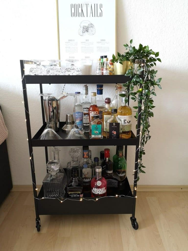 Black three-tier bar cart with a trailing vine plant, cocktail print poster, fairy lights, and a full collection of spirits and decanters