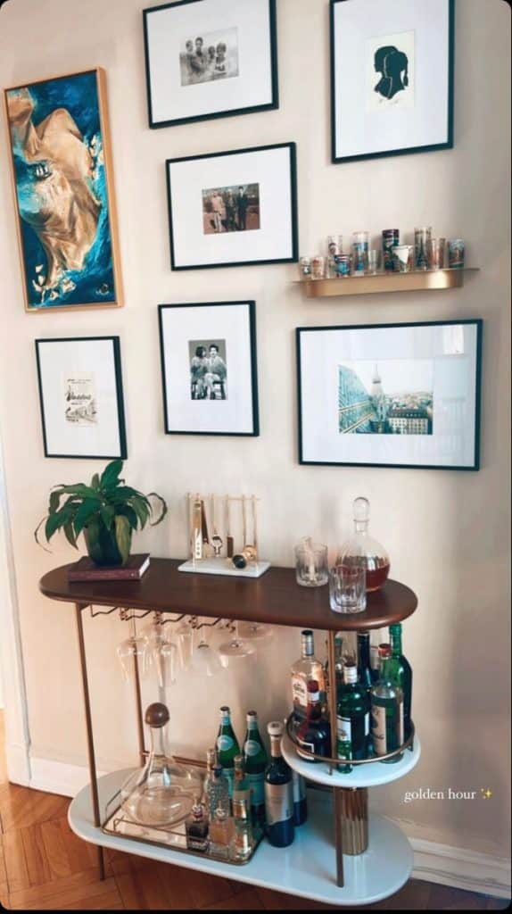 Slim walnut and gold bar cart styled with a decanter, gold bar tools, and hanging wine glasses beneath an eclectic gallery wall of black and white photos and a blue abstract painting