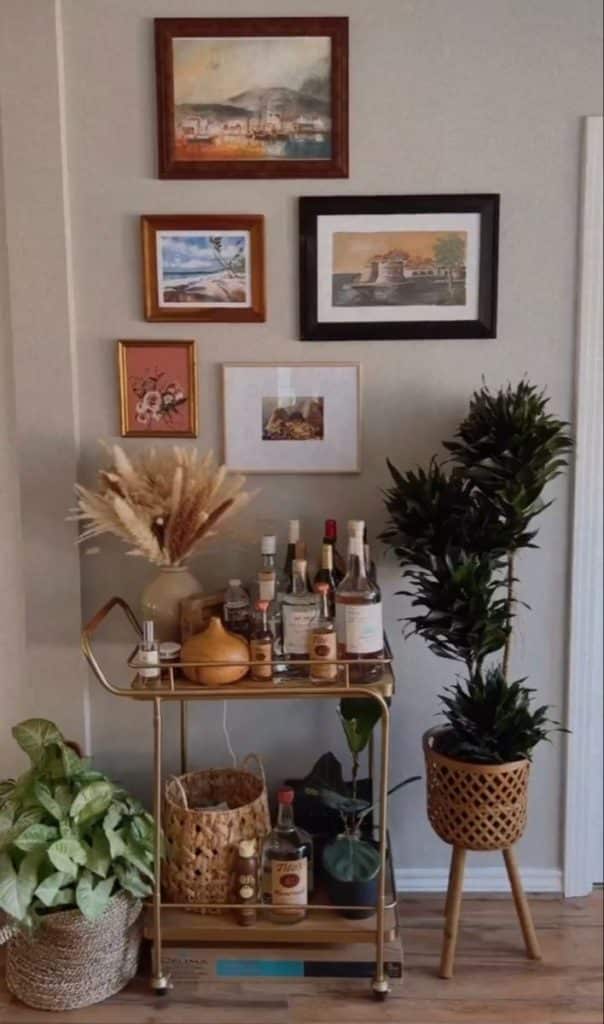 Gold two-tier bar cart styled with pampas grass, a wicker basket, and spirits beneath a collection of thrifted landscape paintings with greenery on either side