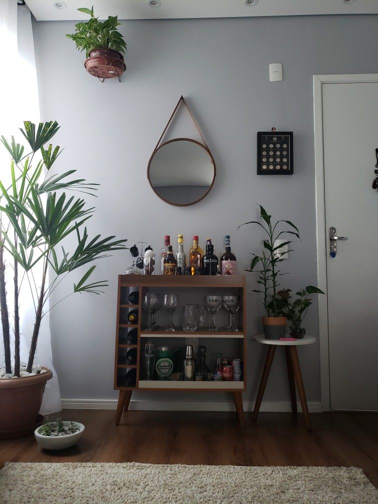 Mid-century walnut drinks cabinet with glass front, integrated wine rack, a collection of spirits on top, and a round leather strap mirror above against a grey wall