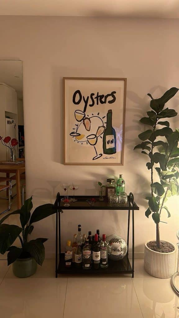 Black two-tier metal bar cart with an oysters and wine art print above, a disco ball on the lower shelf, and tropical plants on either side