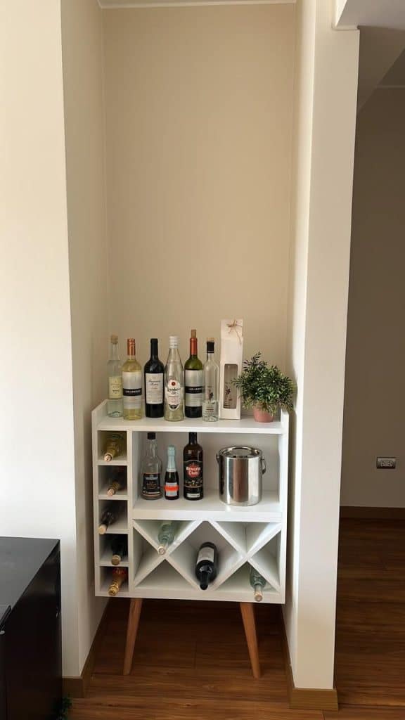 White bar shelving unit with diamond wine storage at the base, upright bottles on top, and a small potted plant, set inside a neutral alcove