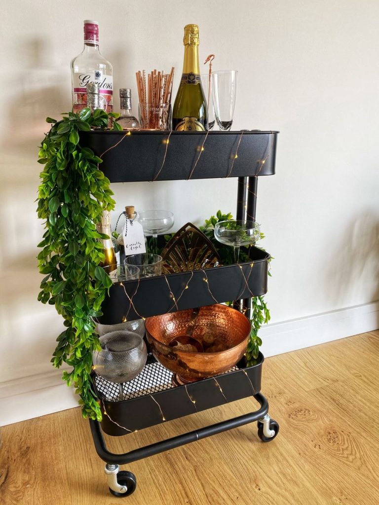 Black three-tier IKEA-style cart draped with a green leaf garland and fairy lights, styled with copper accessories, champagne, and glassware