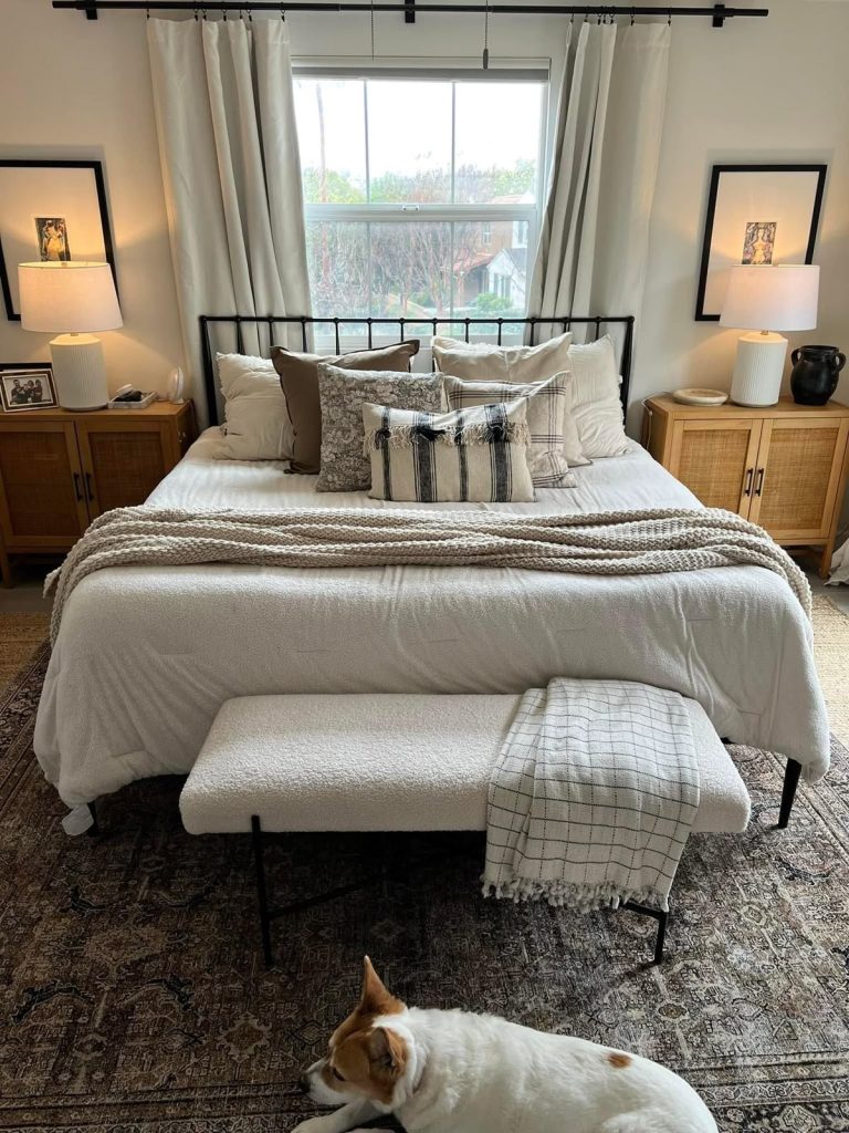 Black iron bed frame with white bedding, a chunky knit throw, and layered neutral and plaid cushions in front of a window with white curtains on a black rod, flanked by rattan cane nightstands with white ceramic lamps and black-framed art, with a woven bench at the foot and a vintage-style area rug