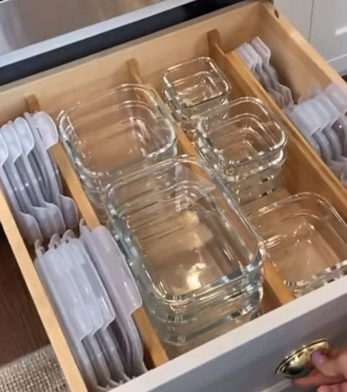 A wide kitchen drawer with wooden dividers on each side holding clear glass food storage containers upright with grey lids sorted behind them.