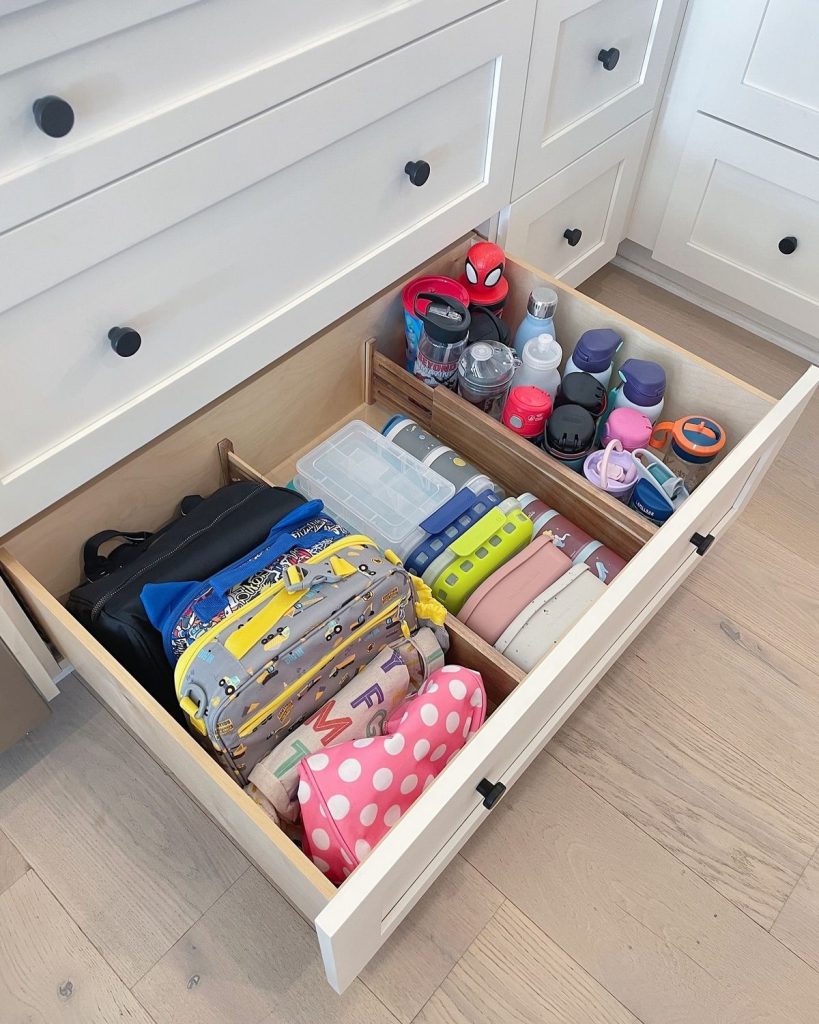 A deep lower kitchen drawer with two sections, one holding children's lunch bags and lunchboxes and the other holding kids water bottles and drink cups sorted upright.