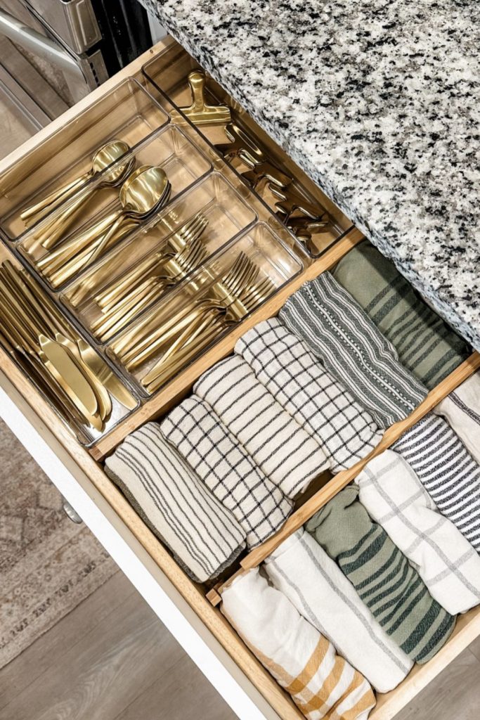 A wide kitchen drawer with a clear acrylic organizer holding sorted gold cutlery on one side and neatly folded striped tea towels stacked on their sides on the other.