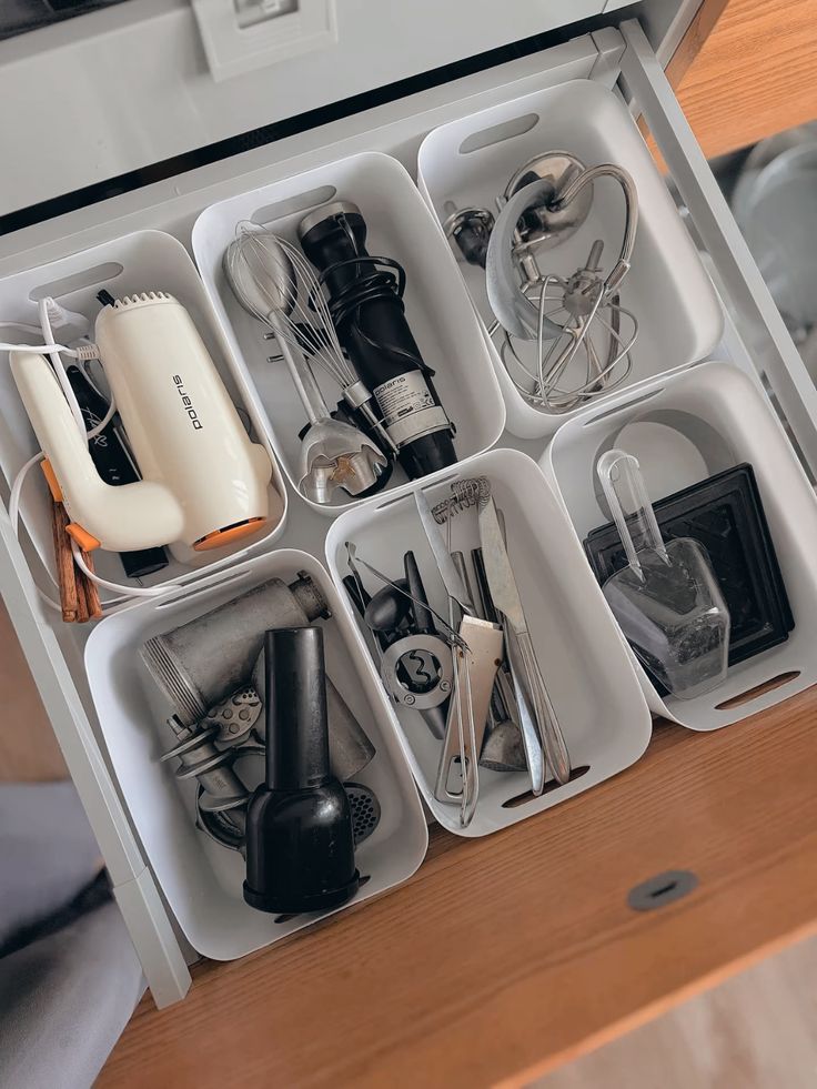 An overhead view of a kitchen drawer with white rectangular bins holding sorted small appliance parts, kitchen tools, and miscellaneous gadgets.