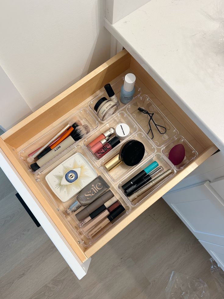 A bathroom drawer with clear acrylic bins holding a curated everyday makeup collection including brushes, lip products, a compact powder, and a mascara in a simple organized layout.