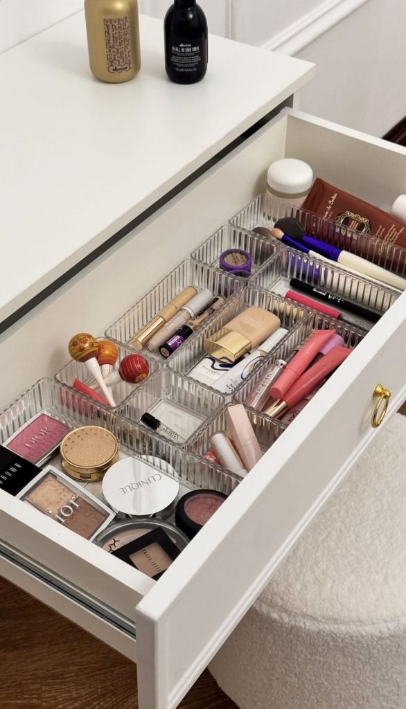 A white vanity drawer organized with ribbed clear acrylic bins holding sorted makeup products including blushes, highlighters, concealers, lip products, and brushes.