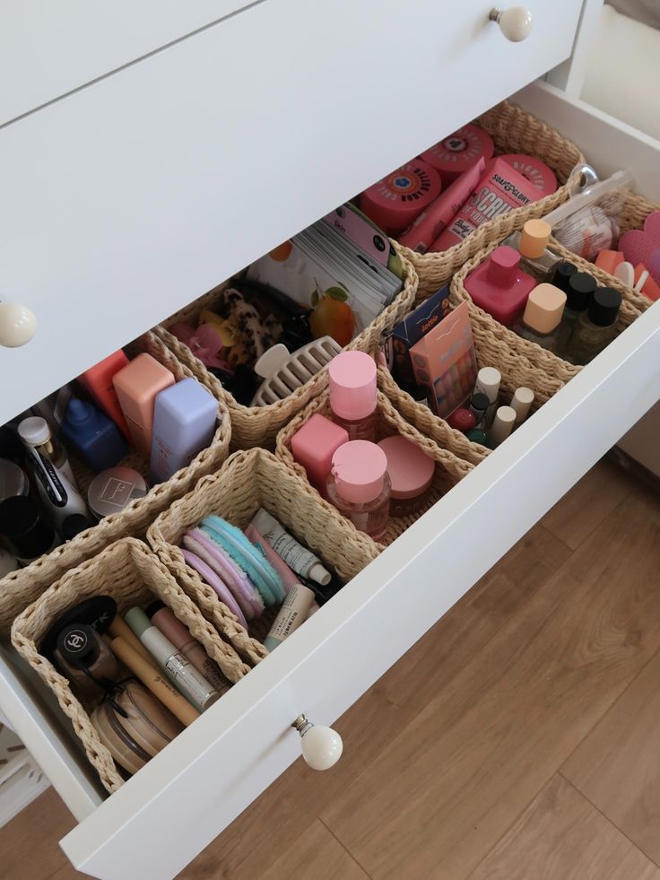 A wide white drawer organized with woven rattan baskets each holding a different category of beauty products including skincare, body products, nail tools, and makeup.