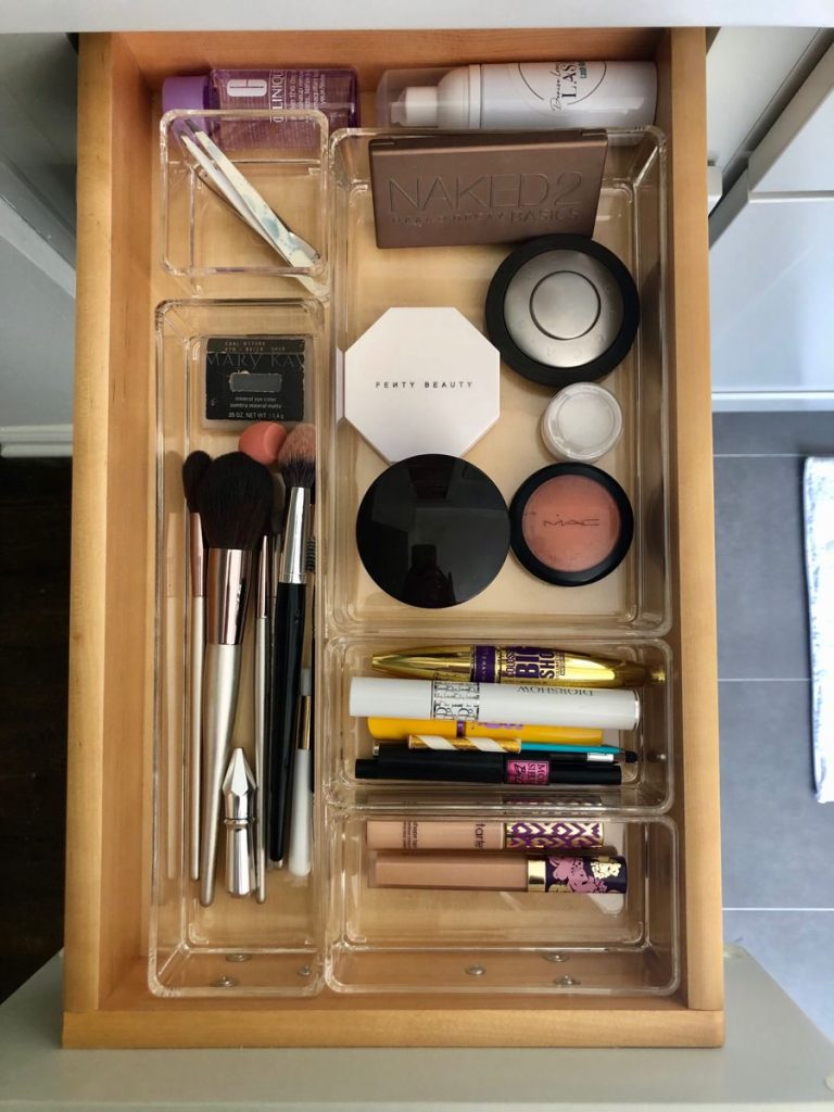 A wooden-based bathroom drawer with clear acrylic bins holding a small organized makeup collection including makeup brushes, face powders, mascaras, and concealers.