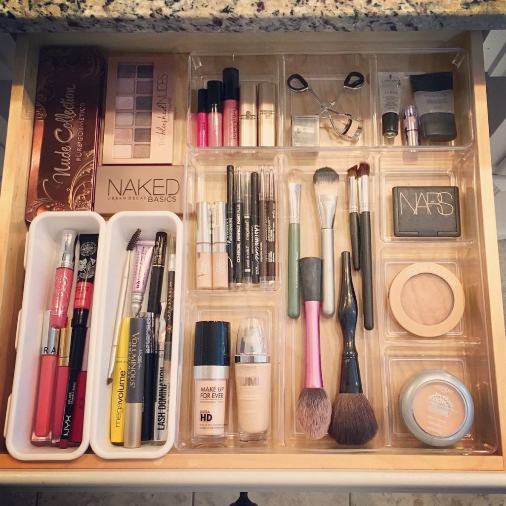 A wooden bathroom drawer organized with a white tray and clear acrylic bins holding eyeshadow palettes, foundations, mascaras, brushes, lip products, and face powders in a full sorted collection.