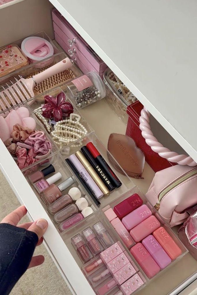 A white vanity drawer organized with clear acrylic bins holding a curated pink-toned makeup collection including pink lip products, nail polishes, mascaras, hair clips, scrunchies, and accessories.
