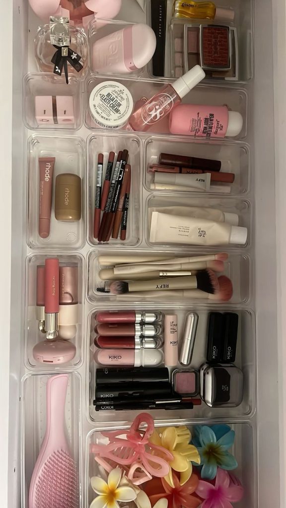 An overhead view of a vanity drawer covered end to end in a grid of clear acrylic bins holding a full sorted makeup collection including lip products, pencils, brushes, mascaras, foundations, and hair accessories.