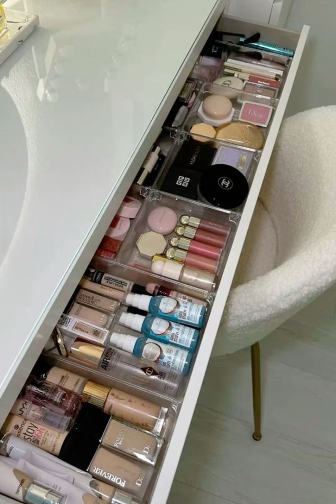 A deep vanity drawer with a full clear acrylic organizer holding sorted foundation bottles, powder compacts, blushes, and lip products in separate compartments.