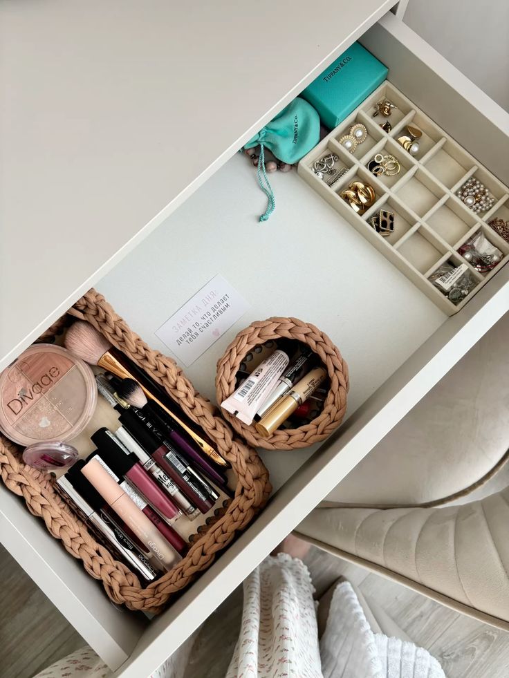 A white vanity drawer with braided baskets holding everyday makeup products and brushes beside a cream divided jewelry tray holding gold earrings, rings, and small accessories.