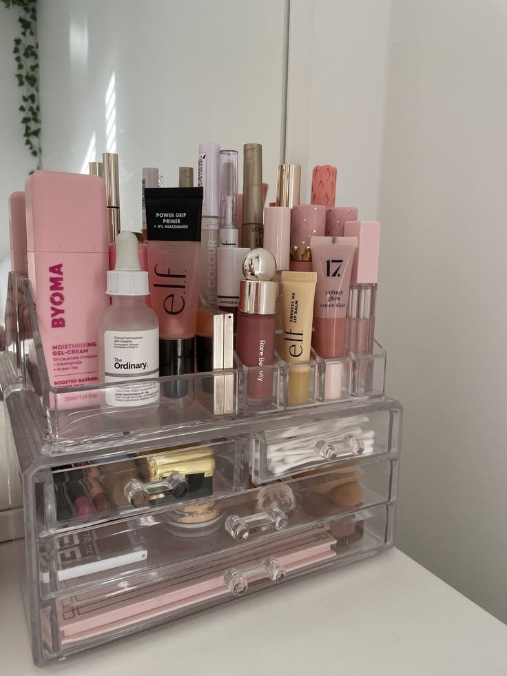A clear acrylic countertop makeup organizer with open slots on top holding skincare and makeup products upright and three small pull-out drawers below holding additional beauty items.