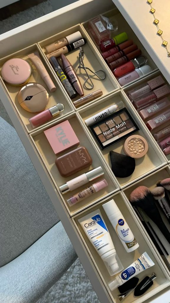 An overhead view of a vanity drawer fitted with a grid of cream-colored square compartments each holding a different category of makeup product including powders, lip glosses, blushes, and brushes.