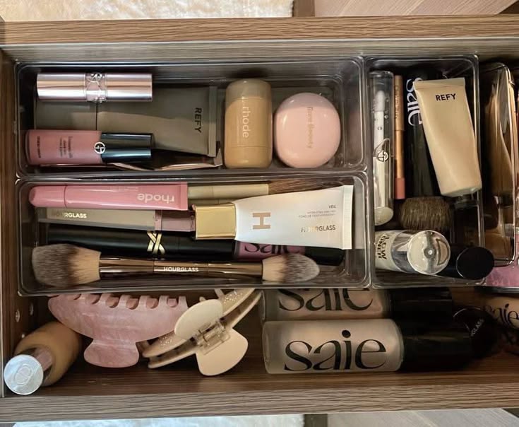 An overhead view of a bathroom drawer with clear acrylic bins holding a curated selection of makeup products including foundations, lip products, face powders, brushes, and a beauty blender.