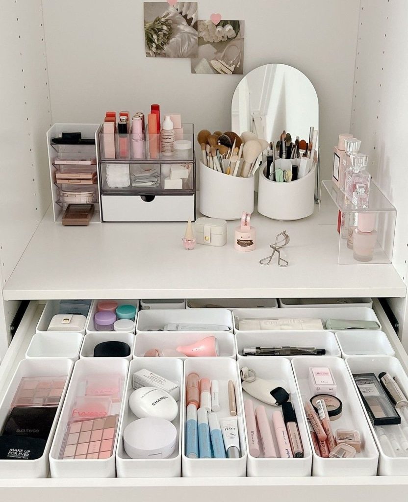 A wide vanity drawer organized with individual white rectangular bins in various sizes holding sorted makeup products including palettes, compacts, lip products, and face tools beside a styled white countertop with brush holders and a mirror.