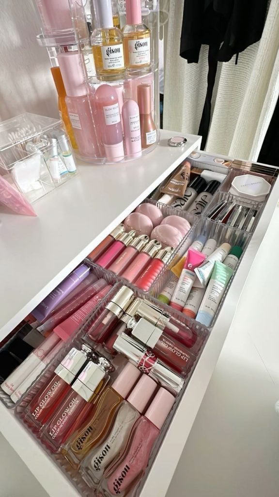 A wide vanity drawer filled with clear acrylic bins holding a large sorted collection of lip glosses, lip oils, lip liners, and lipsticks organized by product type.
