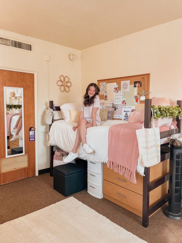 Pink dorm room with a cork board headboard wall, pink throw, and pom pom bedding