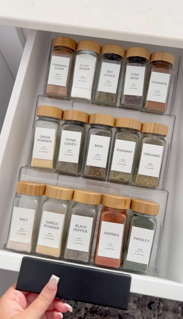 Kitchen drawer with rows of square glass spice jars topped with bamboo lids and clean white printed labels, held in a clear tiered organiser.