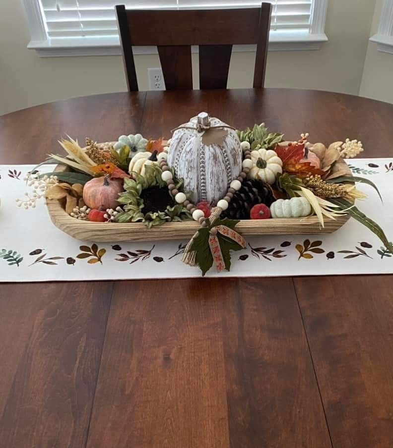Earthy fall dough bowl centerpiece with a large whitewashed carved pumpkin, wood bead garland, mini pumpkins, and fall leaves on a leaf print runner.