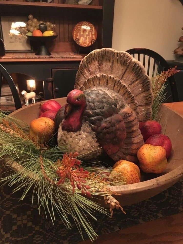 Primitive fall dough bowl centerpiece with a turkey figurine, faux apples and pears, pine greenery, and wheat on a country table.