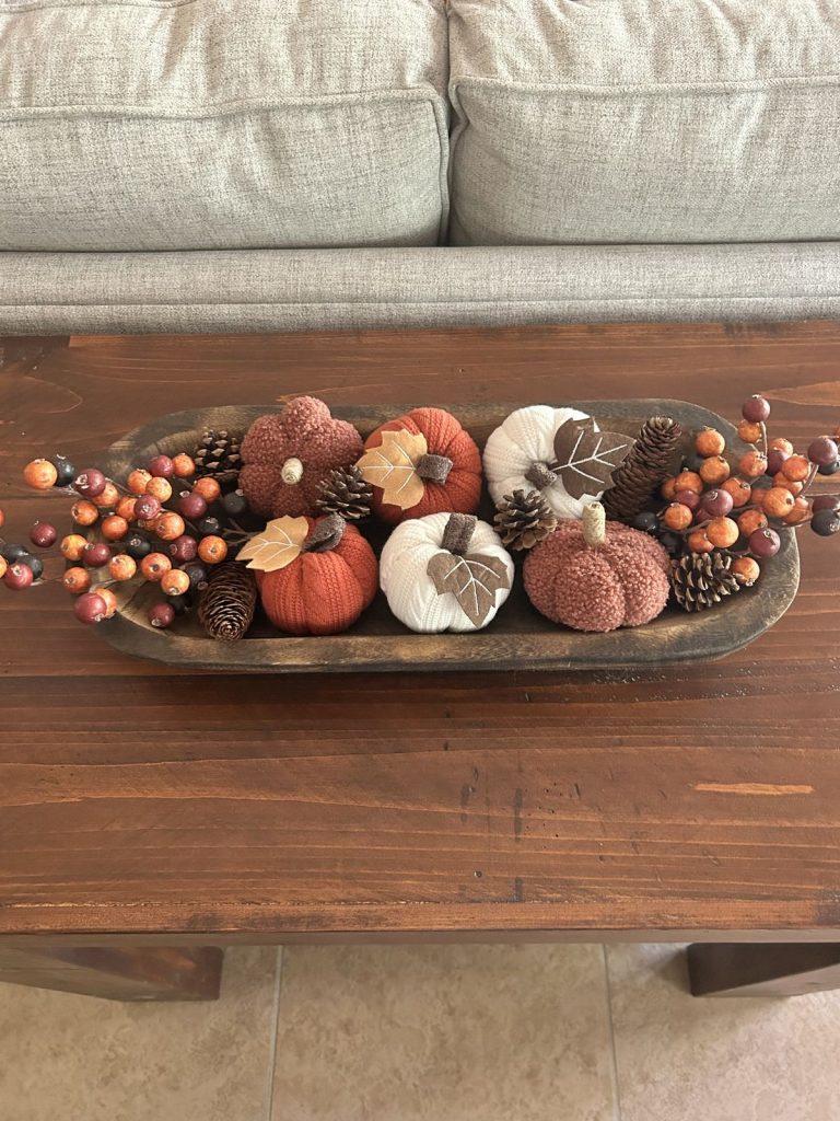 Cozy fall dough bowl filled with rust, orange, and white knit pumpkins, felt leaves, pinecones, and berry sprigs on a wood coffee table.