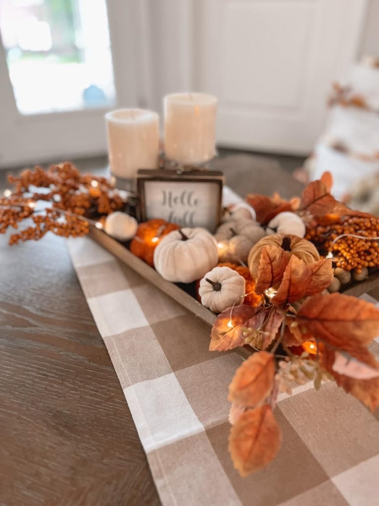 Warm fall dough bowl with a hello fall sign, white and orange velvet pumpkins, cream pillar candles, orange leaves, and fairy lights on a buffalo check runner.