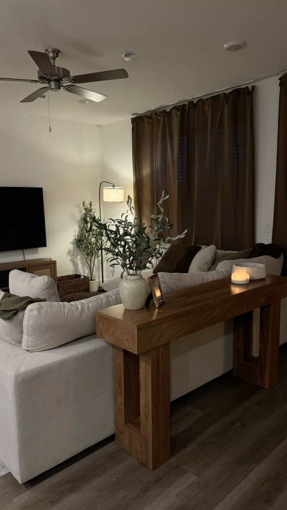 Evening living room with a cream sofa, chunky wooden console table behind it holding a vase of olive branches and a candle, brown curtains, floor lamp, and a wall mounted TV.