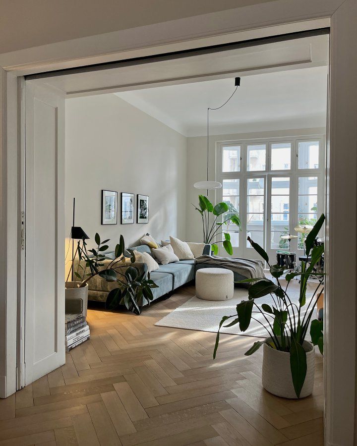 Scandinavian living room seen through open double doors, with herringbone wood floors, a sage blue sofa, three framed prints, several large plants, and a small white pouf.